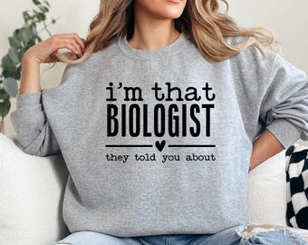Puede incluir: Sudadera gris claro con el texto negro "I'm that BIOLOGIST they told you about". La palabra "BIOLOGIST" está en negrita y en grande. Un pequeño corazón está entre las palabras "BIOLOGIST" y "they".
