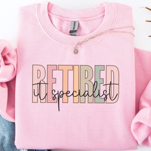 May include: A light pink long-sleeve crewneck sweatshirt featuring the text "RETIRED" in large, blocky, multi-colored letters with the word "it specialist" written in a black cursive script font layered over the center of the design.