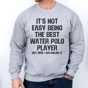 May include: A gray sweatshirt with the text "IT'S NOT EASY BEING THE BEST WATER POLO PLAYER BUT HERE I AM NAILING IT" in black. The sweatshirt is a crew neck style.