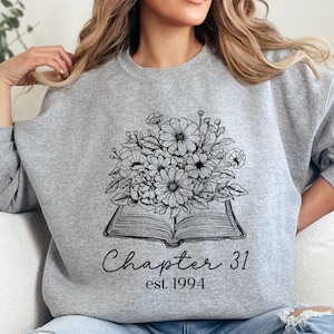 May include: A light gray sweatshirt featuring a black and white floral design atop an open book. The text "Chapter 31 est. 1994" is printed below the book. The sweatshirt is a crew neck style.