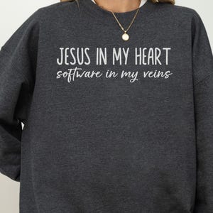 Könnte beinhalten: Ein dunkelgraues Sweatshirt mit Rundhalsausschnitt und dem weißen Aufdruck 'JESUS IN MY HEART' in einer serifenlosen Schrift, gefolgt von 'software in my veins' in einer weißen Schreibschrift auf der Brust.
