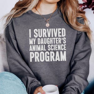 May include: Dark gray sweatshirt with the text "I SURVIVED MY DAUGHTER'S ANIMAL SCIENCE PROGRAM" in white block letters. The sweatshirt is a crew neck style and is being worn by a person.