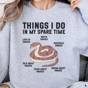 May include: A light grey crewneck sweatshirt featuring a graphic of a coiled brown snake. The text reads: THINGS I DO IN MY SPARE TIME, with labels surrounding the snake: LOOK AT SNAKES, WATCH SNAKES, RESEARCH SNAKES, DREAM ABOUT SNAKES, THINK ABOUT SNAKES, TALK ABOUT SNAKES.