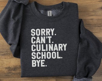 May include: Dark gray sweatshirt with the text "SORRY. CAN'T. CULINARY SCHOOL. BYE." in white block letters. The sweatshirt is folded on a wooden surface.