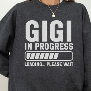 May include: A dark charcoal gray crewneck sweatshirt featuring white screen-printed text that reads GIGI IN PROGRESS above a loading bar graphic and the text LOADING... PLEASE WAIT. The garment has long sleeves and a ribbed collar neckline.