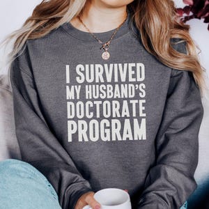 Könnte beinhalten: Dunkelgraues Sweatshirt mit dem weißen Text "I SURVIVED MY HUSBAND'S DOCTORATE PROGRAM". Die Person trägt das Sweatshirt und hält eine weiße Tasse. Das Thema ist akademischer Erfolg.