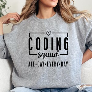 以下が含まれることがあります： 「CODING squad」の文字が黒の太字でプリントされたライトグレーのスウェットシャツ。 「CODING」の文字の上にはハートマークが描かれています。 下には「ALL DAY EVERY DAY」の文字が小さく黒でプリントされています。 カジュアルなスタイルです。