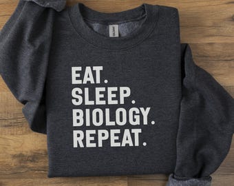 Puede incluir: Sudadera gris oscuro con las palabras "EAT. SLEEP. BIOLOGY. REPEAT." en letras blancas. La sudadera está doblada sobre una superficie de madera.