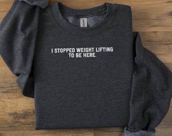 May include: Dark gray sweatshirt with the text "I STOPPED WEIGHT LIFTING TO BE HERE" in white. The sweatshirt is laid flat on a wooden surface. The sleeves are folded over.