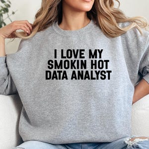 Può includere: Una felpa grigio chiaro con la scritta "I LOVE MY SMOKIN HOT DATA ANALYST" in lettere nere. La maglia a girocollo a maniche lunghe è realizzata in un materiale morbido.