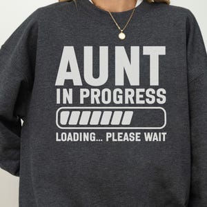 May include: A dark charcoal grey crewneck sweatshirt featuring the white text AUNT IN PROGRESS above a loading bar graphic and the text LOADING... PLEASE WAIT. The design is centred on the chest of the long-sleeved pullover garment.