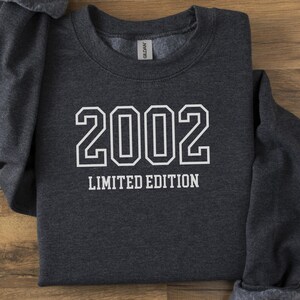 Puede incluir: Una sudadera gris oscuro con el año "2002" en letras blancas grandes y las palabras "LIMITED EDITION" debajo. La sudadera está doblada sobre una superficie de madera.