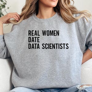 Puede incluir: Una sudadera gris jaspeada con el texto negro "REAL WOMEN DATE DATA SCIENTISTS". La sudadera de cuello redondo está hecha de un material suave. La imagen muestra un primer plano de la parte superior del cuerpo.