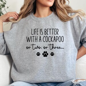 May include: A light gray sweatshirt with the text "LIFE IS BETTER WITH A COCKAPOO or two, or three..." in black. Below the text are three black paw prints. The sweatshirt is a crew neck style.