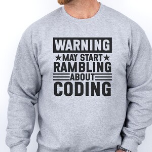 Op de afbeelding: Een lichtgrijze sweater met ronde hals en zwarte tekst die luidt WARNING MAY START RAMBLING ABOUT CODING, met sterren naast het woord MAY en horizontale lijnen die de woorden RAMBLING, ABOUT en CODING scheiden.