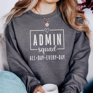 May include: A dark gray sweatshirt with the text "ADMIN squad ALL-DAY EVERY-DAY" in white. The design includes a heart symbol above the word "squad". The sweatshirt is a crew neck style.