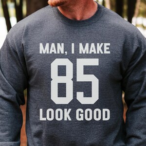 Puede incluir: Una sudadera de cuello redondo en color gris jaspeado oscuro con el texto blanco "MAN, I MAKE 85 LOOK GOOD" impreso en la parte delantera. La prenda tiene mangas largas, cuello acanalado y un corte estándar con letras grandes y llamativas.
