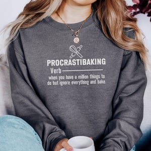 May include: Dark gray sweatshirt with the text "PROCRASTIBAKING" in white, along with a definition and a graphic of a whisk and rolling pin. The sweatshirt is a comfortable, casual style.