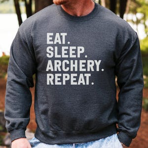 May include: Dark gray sweatshirt with the text "EAT. SLEEP. ARCHERY. REPEAT." in white block letters. The sweatshirt has long sleeves and a crew neck. The background is a blurred outdoor scene with trees.