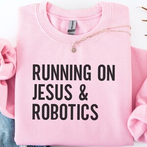 Puede incluir: Una sudadera rosa claro con el texto "RUNNING ON JESUS & ROBOTICS" en letras negras en negrita. La sudadera está doblada, con las mangas y el cuello visibles. Un delicado collar con un pequeño colgante descansa sobre la tela.