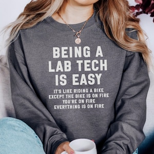 May include: Dark gray sweatshirt with the text "BEING A LAB TECH IS EASY" in white. Below the text is the phrase "IT'S LIKE RIDING A BIKE EXCEPT THE BIKE IS ON FIRE YOU'RE ON FIRE EVERYTHING IS ON FIRE". The sweatshirt is a casual style.