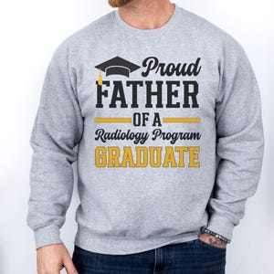 May include: A light gray sweatshirt with the text "Proud Father of a Radiology Program Graduate" in black and gold. The design includes a graduation cap graphic. The sweatshirt is a casual garment.