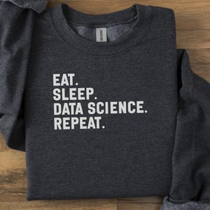 May include: A dark grey sweatshirt featuring the white text "EAT. SLEEP. DATA SCIENCE. REPEAT." The garment is folded on a wooden surface.