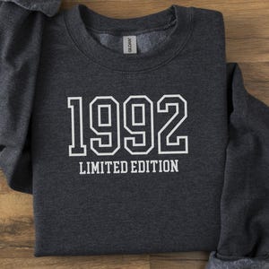 May include: A dark gray sweatshirt with the year "1992" printed in white block letters, with "LIMITED EDITION" below. The sweatshirt is folded on a wooden surface.