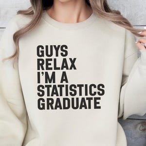 Op de afbeelding: Een beige sweater met ronde hals en zwarte tekst op de voorzijde die luidt: GUYS RELAX I'M A STATISTICS GRADUATE. Het kledingstuk heeft lange mouwen, een geribbelde halslijn en een standaard pasvorm met een duidelijk grafisch ontwerp.
