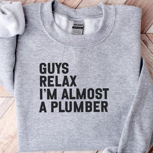 May include: A light gray crewneck sweatshirt featuring the black text "GUYS RELAX I'M ALMOST A PLUMBER" printed on the front. The garment has long sleeves, a ribbed collar, and a Gildan brand label visible on the inside of the neckline.