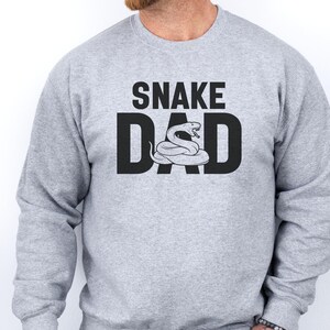 Könnte beinhalten: Ein hellgraues Sweatshirt mit dem Aufdruck "SNAKE DAD" in schwarzen Buchstaben. Das "O" in "DAD" wird durch eine Schlange ersetzt. Das Sweatshirt hat einen Rundhalsausschnitt.