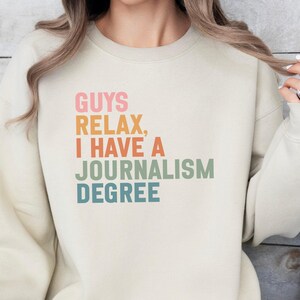 May include: A cream-colored sweatshirt with the text "GUYS RELAX, I HAVE A JOURNALISM DEGREE" in a colorful, stacked font. The sweatshirt has long sleeves and a crew neck.