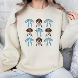 May include: Cream-colored sweatshirt featuring a repeating pattern of brown and white dogs with blue bows. The dogs have a white chest and a brown head with brown spots. The bows are light blue.