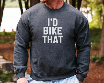 Puede incluir: Sudadera gris oscuro con el texto blanco "I'd Bike That". La sudadera de cuello redondo tiene mangas largas. El fondo es una escena borrosa al aire libre con árboles.