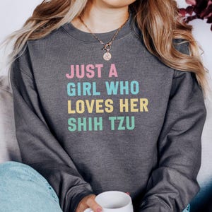 Puede incluir: Sudadera gris oscuro con el texto "JUST A GIRL WHO LOVES HER SHIH TZU" en rosa, azul, amarillo y verde. La sudadera es de cuello redondo y está hecha de un material suave. Una persona lleva la sudadera.