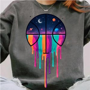 May include: Dark gray sweatshirt featuring a basketball design with a space theme. The basketball graphic has a night sky background with planets and a crescent moon. Colorful drips of orange, pink, teal, and purple flow from the bottom of the basketball.
