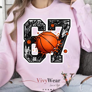 Könnte beinhalten: Hellrosa Sweatshirt mit Basketball-Design. Das Design zeigt die Zahl "87" in schwarzer, gekritzelter Schrift, mit einem orangefarbenen Basketball in der Mitte und kleineren Bällen darum herum. Der Markenname "VivyWear" ist unten aufgedruckt.