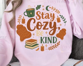 Może przedstawiać: Jasnoróżowa bluza z nadrukiem graficznym. Projekt zawiera słowa "Stay Cozy, Be Kind" w brązowych i pomarańczowych literach, a także ilustracje kubka, koca, książek i świec.