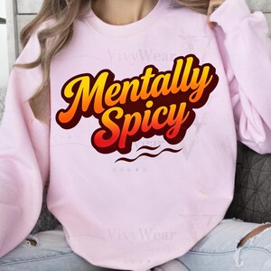 Op de afbeelding: Een lichtroze sweatshirt met de tekst "Mentally Spicy" in een gedurfde, gestileerde lettertype. De tekst is in tinten geel, oranje en rood, met een donkere omtrek. Twee golvende lijnen staan eronder.