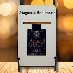 May include: A magnetic bookmark with the text "Magnetic Bookmark" at the top. The bookmark features a movie poster from the film "SAW". The bookmark is held by a black metal stand on a wooden surface.