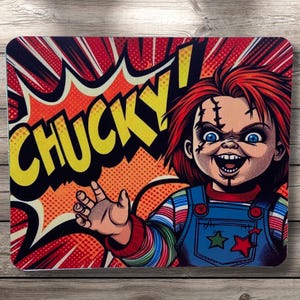 May include: A rectangular mousepad featuring a cartoon image of Chucky from the horror film series. The image shows Chucky with red hair, blue eyes, and a stitched face, set against a comic book-style background with the word "CHUCKY!"