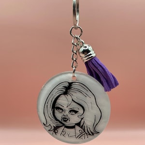 May include: A round keychain featuring a black and white illustration of a doll's face. The keychain has a silver chain and a purple tassel. The background is a soft pink.