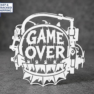 May include: A white vinyl decal featuring the text GAME OVER in a stylized, dripping font. The design is enclosed within a circular frame adorned with mechanical, gear-like components and a jagged, spiked border along the bottom edge.