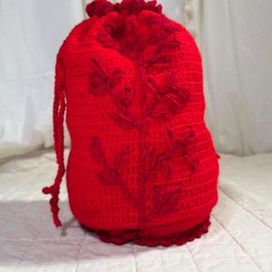 May include: A handmade, bright red crochet drawstring bag. The bag features a floral design and a scalloped edge. The drawstring is made of the same red yarn. The bag is likely used for storage or as a gift bag.