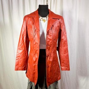 May include: A vintage, red leather jacket with a collared neckline and button-up closure. The jacket features two front pockets and a tailored fit. The jacket is displayed on a mannequin, paired with a white top and a black skirt.