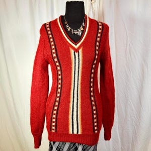 Puede incluir: Un suéter rojo con cuello en V con un diseño geométrico. El suéter presenta un patrón de rayas verticales blancas, negras y beige en la parte delantera y detalles punteados en los laterales. Un collar plateado es visible en el escote.