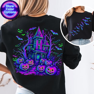 May include: Black sweatshirt with a Halloween-themed design. The back features a haunted house, pumpkins, bats, and trees in neon green, purple, and blue. The front has a design of bats. The words "More Color Options" are in a circle.