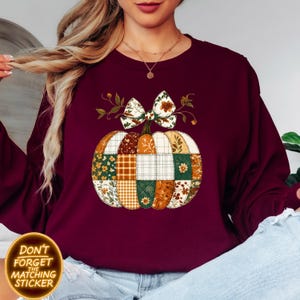 K&ouml;nnte beinhalten: Burgunderfarbenes Sweatshirt mit einem Patchwork-K&uuml;rbis-Design, einer Schleife und floralen Akzenten. Der K&uuml;rbis besteht aus verschiedenen gemusterten Stoffen in Orange, Gr&uuml;n und Creme. Der Text "DON'T FORGET THE MATCHING STICKER" befindet sich in einem goldenen Kreis.