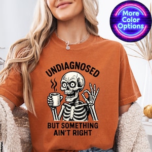 May include: Burnt orange t-shirt with a skeleton graphic holding a coffee mug and giving an 'okay' hand gesture. The text on the shirt reads "UNDIAGNOSED BUT SOMETHING AIN'T RIGHT". A neon sign in the upper right corner says "More Color Options".