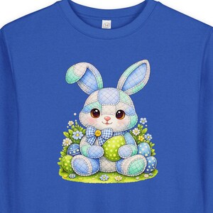 Könnte beinhalten: Blaues Sweatshirt mit einem Osterhasen-Motiv. Der Hase ist im Patchwork-Stil gehalten und hält ein grünes Ei, umgeben von Blumen und Ostereiern. Der Hase hat blaue und grüne Ohren und eine blaue Fliege.
